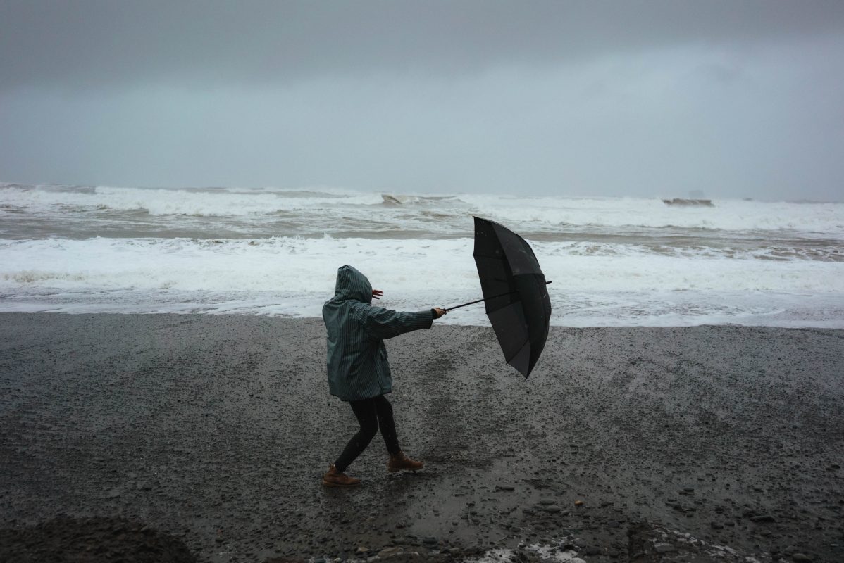 unbekannte Person am Strand mit Schirm, müht sich gegen den Wind